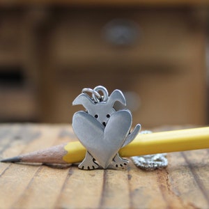 May include: Silver charm of a dog holding a heart in its paws. The charm is on a pencil.