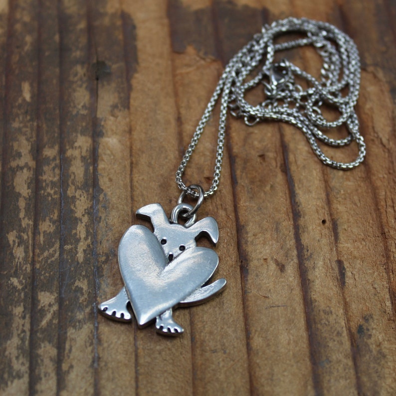 May include: A silver necklace with a pendant of a dog holding a heart. The pendant is made of pewter and has a detailed design.