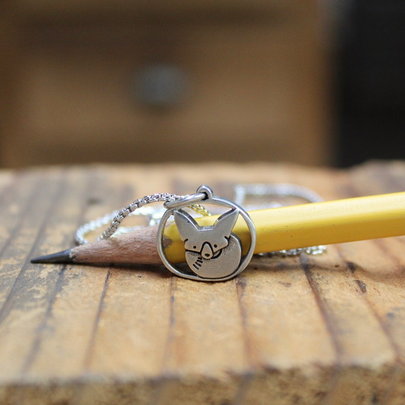 May include: A silver necklace with a pendant featuring a fox's face. The pendant is round and has a detailed design. The necklace is resting on a wooden surface with a yellow pencil.