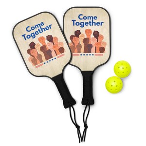 May include: Two wooden pickleball paddles with the text "Come Together" and a graphic of raised fists in various skin tones. The paddles have black handles and are paired with two yellow pickleballs. The paddles are designed for playing pickleball.