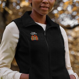 May include: Black fleece vest with a full-length zipper and a stand-up collar. The vest features the embroidered text "Come Together" above a graphic of raised hands in various colors. The model is wearing a white long-sleeve shirt underneath.