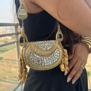 Op de afbeelding: Een goudkleurige, halfronde handtas met ingewikkeld borduurwerk, kleine vierkante spiegelversieringen en bungelende goudkleurige muntbedels. De tas heeft een lange, decoratieve schouderband die is versierd met extra spiegelaccenten.