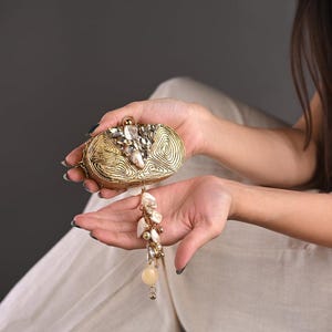 May include: A gold-colored clutch with a textured, spiral design. The top of the clutch is embellished with clear and iridescent stones. A decorative tassel of pearls and beads hangs from the bottom. The clutch is held in open hands.