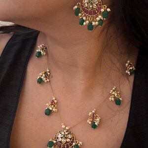 May include: A gold-toned jewelry set featuring a necklace and earrings. The necklace has a pendant with red and clear stones, green beads, and white pearls. The earrings have a similar design. The jewelry is worn against a dark background.