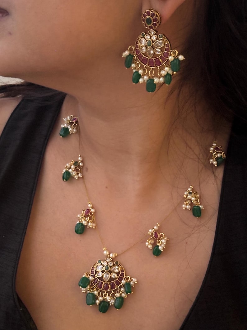 May include: A gold-toned jewelry set featuring a necklace and earrings. The necklace has a pendant with red and clear stones, green beads, and white pearls. The earrings have a similar design. The jewelry is worn against a dark background.