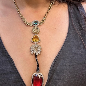 May include: A gold necklace with a series of decorative pendants. The necklace features a large, red gemstone pendant at the bottom, with a black and gold stem. Above are a flower, a yellow gem, a lion's head, and a green gem.