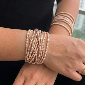 May include: A set of rose gold-toned tennis bracelets featuring multiple rows of small, clear, sparkling stones. The bracelets are worn in a stacked, crisscrossed arrangement on the wrist, with additional matching bracelets on the other arm.