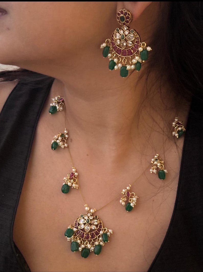 May include: Gold-toned jewelry set featuring a necklace and earrings. The necklace has a pendant with green and red gemstones, and the earrings have a similar design. The set includes small pearls and green teardrop-shaped stones.