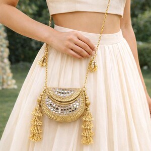 May include: Gold-coloured, half-moon shaped handbag with a chain strap. The bag is embellished with small mirrored squares and tassels. The chain and tassels are also gold. The bag is held in front of a cream-coloured outfit.