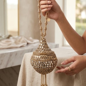 Pode incluir: Uma bolsa dourada ornamentada, com uma alça de contas e um corpo em forma de gota. A bolsa é coberta por pequenos enfeites reflexivos e tem borlas de corrente penduradas com pequenas contas douradas.