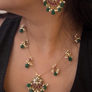 May include: A gold-toned jewelry set featuring a necklace and earrings. The necklace has a pendant with a circular design, adorned with red, green, and white gemstones. The earrings mirror the pendant design. The set includes small, dangling green beads.