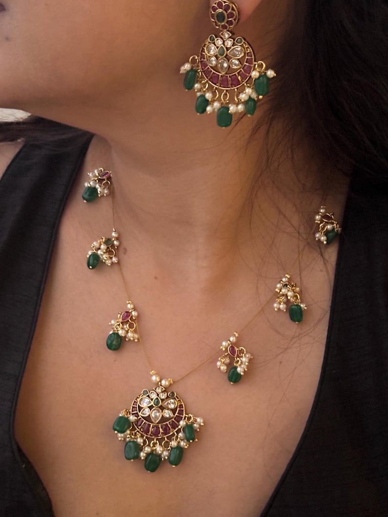 May include: A gold-toned jewelry set featuring a necklace and earrings. The necklace has a pendant with a circular design, adorned with red, green, and white gemstones. The earrings mirror the pendant design. The set includes small, dangling green beads.