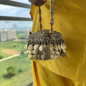 Könnte beinhalten: Eine goldfarbene Metallhandtasche mit quadratischem Körper, verziert mit komplizierten Filigranmustern und einem dichten Saum aus überlappenden, kreisförmigen Metallscheiben. Die Tasche hat einen hohen, schlanken Griff mit segmentierter Perlenstruktur.