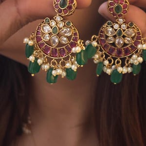 May include: Gold-toned earrings with a floral design. The earrings feature a circular design with red and green gemstones, clear crystals, and dangling green beads and small white pearls. The earrings are held up by a person.