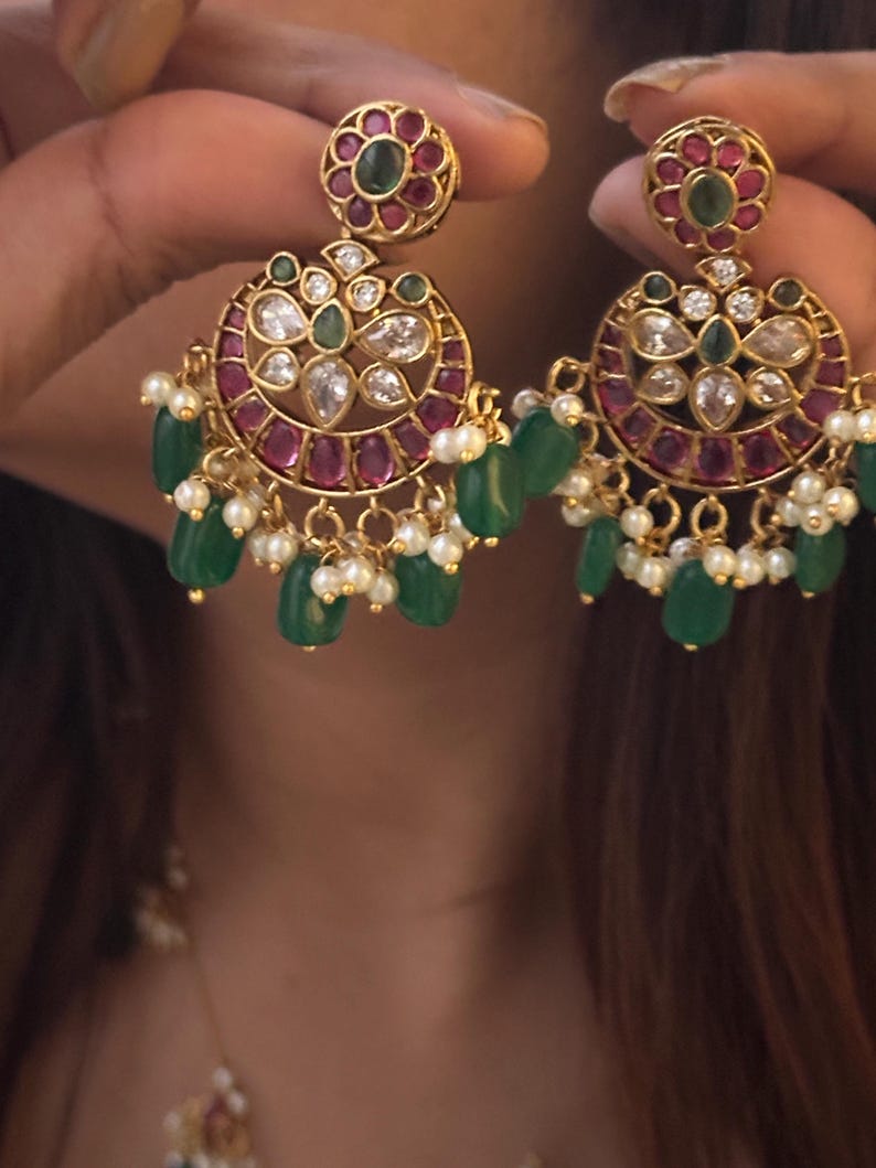 May include: Gold-toned earrings with a floral design. The earrings feature a circular design with red and green gemstones, clear crystals, and dangling green beads and small white pearls. The earrings are held up by a person.