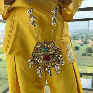 Puede incluir: Un bolso de mano hexagonal de metal dorado con un intrincado patrón en relieve, una piedra ovalada verde central y una piedra ovalada roja más grande. El bolso cuelga de una cadena adornada con racimos de pequeñas conchas blancas y cuentas doradas.