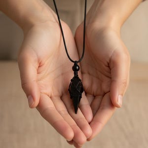 May include: A black pendant necklace featuring a geometric crystal encased in a woven black cord. The necklace has a black bead and is suspended from a black cord. The pendant is held in open hands.