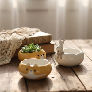 May include: Three ceramic planters on a wooden surface. Two are yellow with white faces, and one is white with a bunny design. One yellow planter contains a green succulent. The planters are decorative.