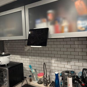 Könnte beinhalten: Ein weißer Tablethalter an einer weiß gefliesten Küchenwand, der ein schwarzes Tablet hält. Die Küche verfügt über ein Edelstahlbecken, einen Toaster und einen Reiskocher. Schränke sind im Hintergrund sichtbar.
