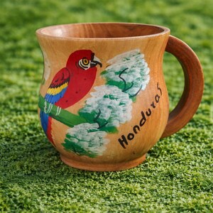 May include: Wooden mug with a painted parrot design. The parrot is red, blue, and yellow, perched on a green branch with white flowers. The word "Honduras" is written on the mug. The mug has a natural wood finish.
