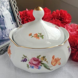 May include: A white porcelain lidded dish with a gold rim and a gold-tipped handle. The dish is decorated with colorful floral patterns, including roses and tulips. The background includes red flowers and a framed black and white photograph.