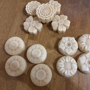 May include: A collection of beige-colored, floral-shaped soaps. The soaps are in various designs, including round sunflower shapes and flower shapes. The soaps are arranged on a wooden surface.