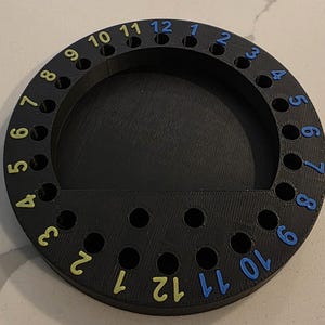 May include: A black, circular organizer with numbered holes. The numbers 1-12 are printed in yellow and blue around the outer edge. The organizer has a semi-circular tray at the bottom and is likely used for storing small items.