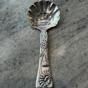 May include: A silver spoon with a scalloped bowl and a floral design on the handle. The handle features detailed carvings of flowers and leaves, adding a decorative touch to the utensil. The spoon is likely made of a metal alloy.