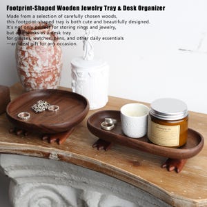 May include: Footprint-shaped wooden jewelry tray and desk organizer. The dark brown trays are made of wood and have a footprint design. One tray holds rings, and the other holds a candle and a jar.