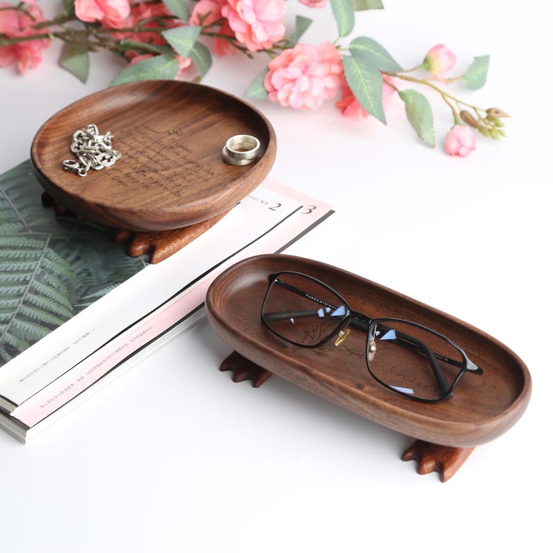 May include: Two wooden trays, one round and one oval, with a dark brown finish. The round tray holds a silver chain and a ring. The oval tray holds a pair of eyeglasses. Both trays have decorative feet.