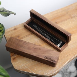 May include: A wooden jewelry box with a hinged lid, open to reveal a silver necklace on a black velvet lining. The box is rectangular and made of dark wood. The closed box has the word "SwalRidge" engraved on the side.