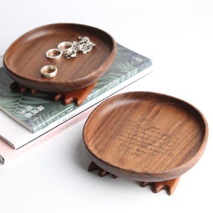 May include: Two wooden jewelry trays with a dark brown finish. One tray holds rings and a necklace. The other tray has an engraved quote. Both trays are on small wooden feet and rest on a book.