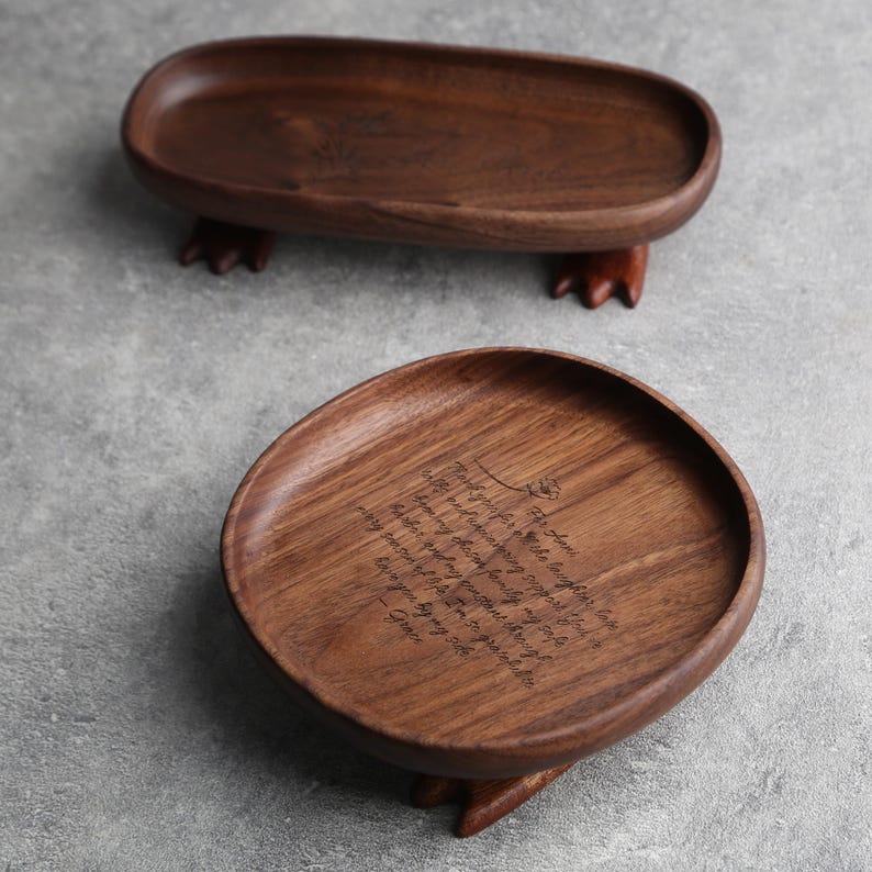 May include: Two handcrafted wooden trays. One is oval-shaped, the other is round with a slightly irregular edge. Both are dark brown and rest on small, carved feet. The round tray has engraved text.