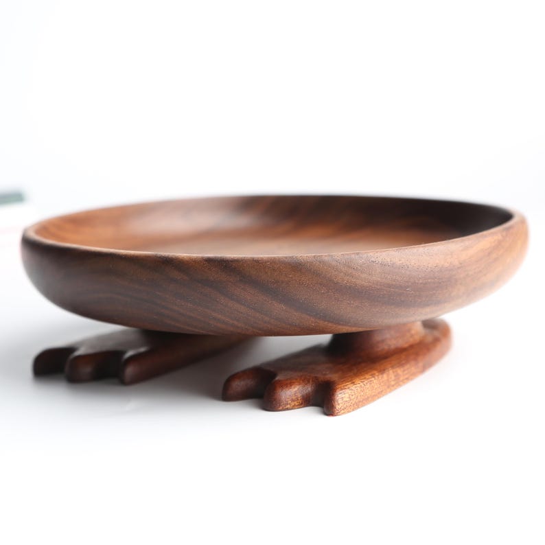May include: A handcrafted wooden bowl with a unique design. The bowl is a warm brown color and is supported by two carved feet. The bowl is perfect for serving snacks or as a decorative piece. The bowl measures approximately 10 inches in diameter.