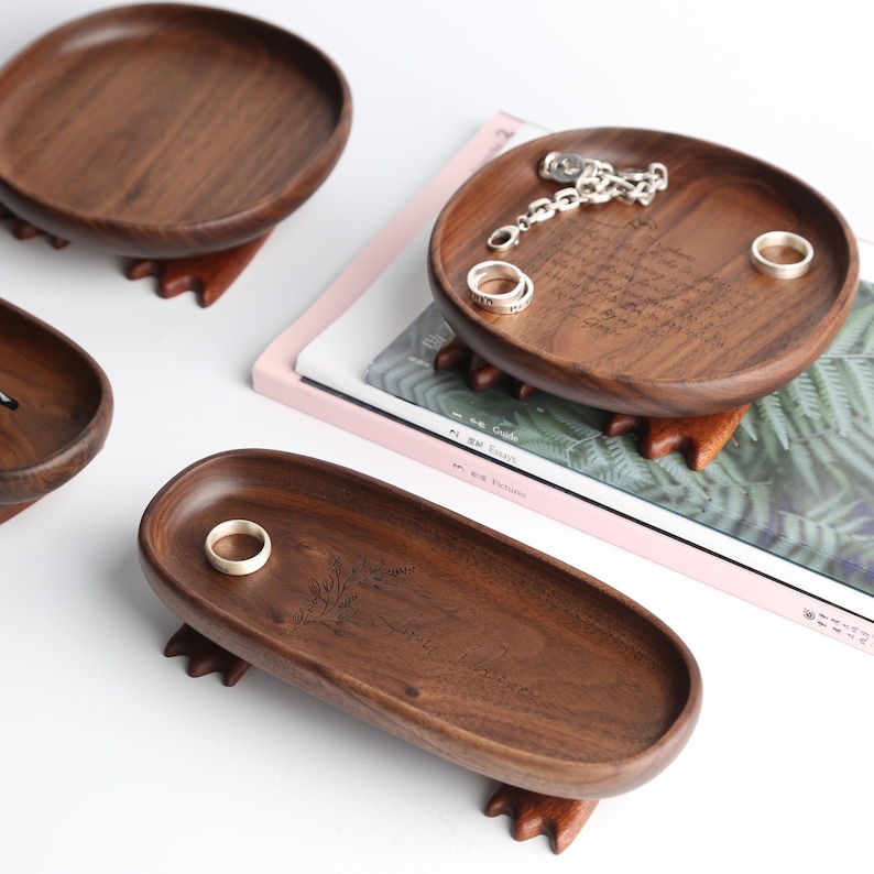 May include: Wooden jewelry trays in various shapes, including round and oval, with a dark brown finish. The trays are elevated on small, claw-like feet. Some trays hold rings and a silver chain. The background is white.