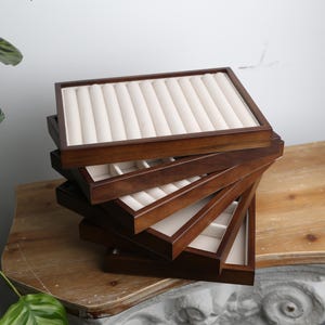 Peut inclure: Une pile de plateaux à bijoux en bois avec une finition marron foncé. Chaque plateau a un intérieur rembourré de couleur crème avec des fentes pour les bagues et autres bijoux. Les plateaux sont disposés de manière légèrement éventail, mettant en valeur leur design.