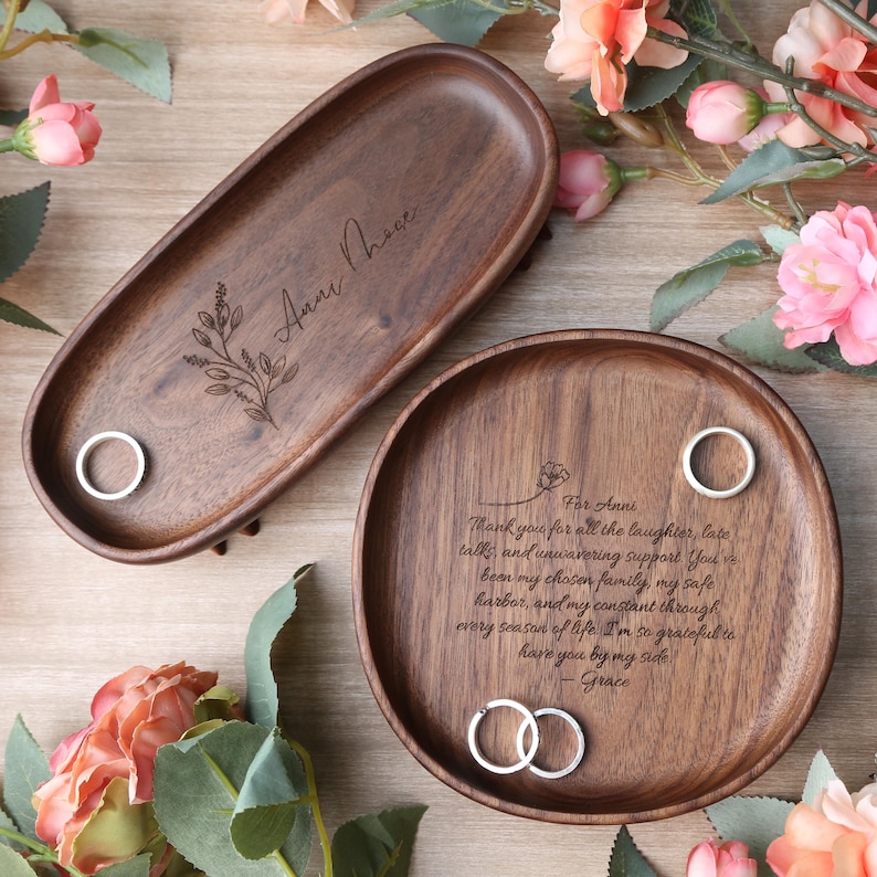 May include: Two wooden trays, one oval and one round, with engraved text and floral designs. The oval tray has the name "Anni Those" and a floral sprig. The round tray has a message and two rings. Both trays have a ring holder.