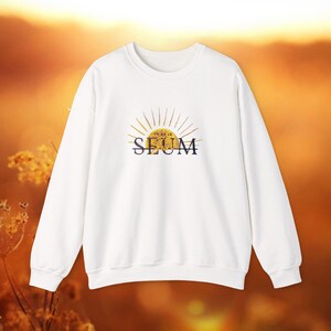 May include: White crew neck sweatshirt with a gold sunburst design and the word "SEUM" in navy blue. The sweatshirt has long sleeves and a relaxed fit, suitable for casual wear.