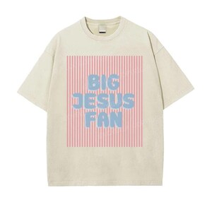 May include: Cream-colored t-shirt with a crew neck. The front features a rectangular design with pink vertical stripes and the words "BIG JESUS FAN" in blue bubble letters.