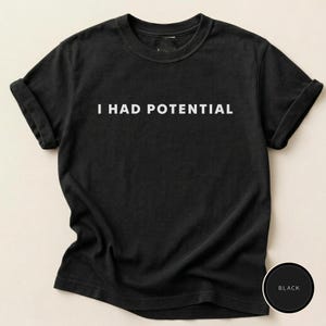 Può includere: T-shirt nera con la scritta bianca "I HAD POTENTIAL". La maglietta ha maniche corte e girocollo. L'immagine include un cerchio nero con la parola "BLACK" in testo bianco.