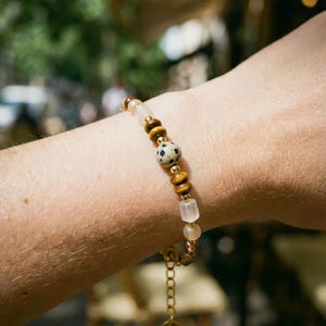Könnte beinhalten: Ein Armband mit einer Mischung aus Natursteinperlen in Braun-, Beige- und Cremetönen. Das Armband enthält eine große, gesprenkelte Perle und ist mit einer goldfarbenen Metallkette und einem kleinen Anhänger versehen.