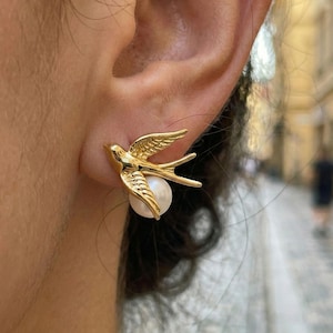 Puede incluir: Pendientes de botón dorados con forma de pájaro y un acento de perla. Los pendientes presentan un diseño detallado de pájaro con alas extendidas y una perla blanca redonda. Los pendientes se llevan en una oreja.