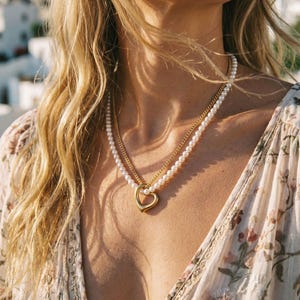 Può includere: Una collana a strati composta da un filo di piccole perle bianche e una sottile catena in metallo dorato. Un ciondolo liscio e lucido a forma di cuore color oro è sospeso al centro dove le due catene si incontrano, poggiando sulla clavicola.
