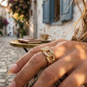 Puede incluir: Un anillo de eslabones dorados que presenta una serie de piedras transparentes de corte baguette incrustadas en la superficie de cada eslabón entrelazado. El anillo tiene un acabado metálico pulido y un diseño de banda ancha y texturizada.