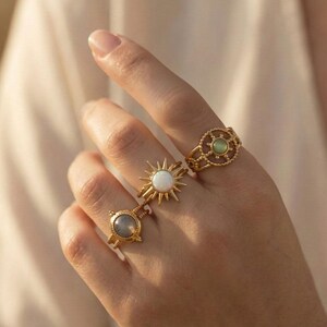 Könnte beinhalten: Eine Hand zeigt drei goldene Ringe. Ein Ring hat ein Sonnenstrahl-Design mit einem weißen Stein in der Mitte. Ein anderer hat einen kreisförmigen grünen Stein, und der dritte hat einen grauen Stein. Alle Ringe haben aufwendige Details.