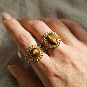 Könnte beinhalten: Zwei goldfarbene Ringe mit Tigeraugensteinen. Ein Ring hat ein Sonnenmuster, der andere einen ovalen Stein mit einem seilartigen Rand. Die Ringe werden an einer Hand getragen.