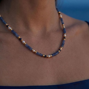 Può includere: Una collana di perline con dettagli blu, bianchi e dorati. La collana presenta piccole perline blu, intervallate da perle bianche e distanziatori color oro. La collana è indossata intorno al collo, con uno sfondo blu scuro.
