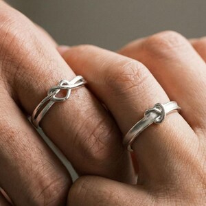 Könnte beinhalten: Zwei silberfarbene Knotenringe werden auf Fingern gezeigt. Die Ringe haben ein schlichtes, elegantes Design, wobei das Metall eine Knotenform bildet. Die Ringe befinden sich an den Fingern einer Person, mit einem neutralen Hintergrund.