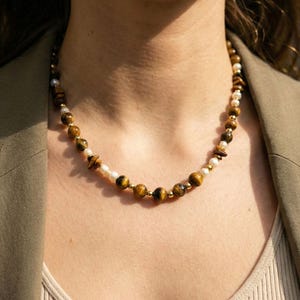Puede incluir: Un collar de cuentas con una alternancia de piedras redondas de ojo de tigre, pequeñas perlas blancas y diminutas cuentas espaciadoras de metal dorado. El collar presenta un patrón repetitivo de piedras marrones y doradas, perlas blancas brillantes y acentos metálicos.