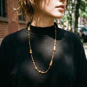 Op de afbeelding: Een lange ketting met een reeks bruine en goudgele tijgeroogkralen, afgewisseld met kleinere, ronde, donkerbruine kralen. Het ontwerp bevat langwerpige, druppelvormige tijgeroogstenen die gelijkmatig over het dunne koord zijn verdeeld.